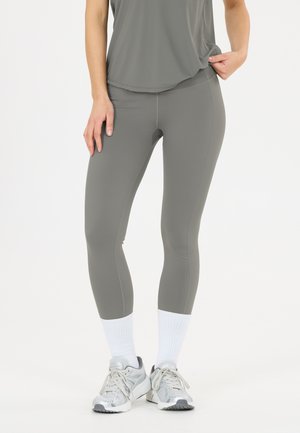 Persoon draagt grijze leggings, bijpassende grijze top, witte sokken en grijze sportschoenen en staat voor een effen lichte achtergrond.