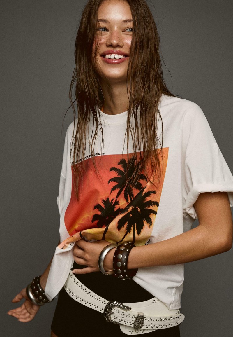 White T-shirt features a sunset graphic with palm trees, paired with a black mini skirt and layered bracelets. Accents include a studded belt.