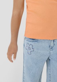 Persona vistiendo jeans azul claro con un pequeño bordado de flores en el bolsillo y una camisa de color durazno, con la mano relajada a un lado.