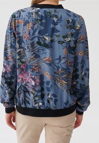 Květinová bomber bunda z modré látky, s barevnými botanickými potisky, černým žebrovaným límcem a manžetami, a elastickým lemem.