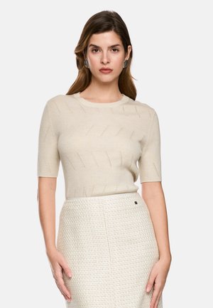Woman with long brown hair wearing a beige short-sleeve knit top and a high-waist textured white skirt against a plain background.