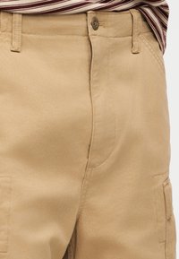 Κοντινή εικόνα από μπεζ παντελόνι cargo με κούμπωμα σε κουμπί και θηλιές για ζώνη, φορεμένο με κόκκινη και λευκή ριγωτή μπλούζα που είναι κλειδωμένη μέσα.