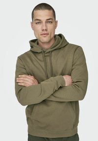 Sudadera verde oliva con un bolsillo canguro en el frente, capucha con cordones, puños acanalados y una textura suave. El modelo cruza los brazos, mostrando la prenda.