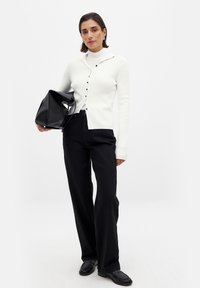 Valge soonikkoes kardigan mustade nööpidega, kokku sobitatud mustade laiade pükstega ja musta kotiga, lihtsa disaini ja tekstuuriga.