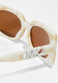 Overdrevne solbriller med en marmoreret ramme i creme og brun plast, brune tonede linser og et metallisk logo på bøllen.