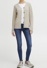 Beżowy dziergany kardigan z teksturowanej tkaniny, z pięcioma brązowymi guzikami, noszony na białej koszulce, zestawiony z niebieskimi obcisłymi dżinsami i jasnoszarymi trampkami.