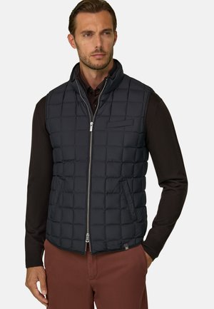Mann trägt eine schwarze, gesteppte ärmellose Weste über einem schwarzen Langarmshirt und braune Hosen, steht mit einer Hand in der Tasche.