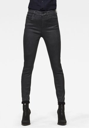 Osoba nosząca dopasowane czarne jeansy z przednimi kieszeniami i czarne botki z zamkami, stojąca na tle jednolitego tła.