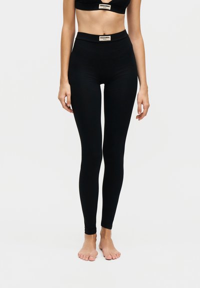 Жена, носеща черни клинове с висока талия и съответстващ къс топ, и двата с бежови лога DSQUARED2 Milano.