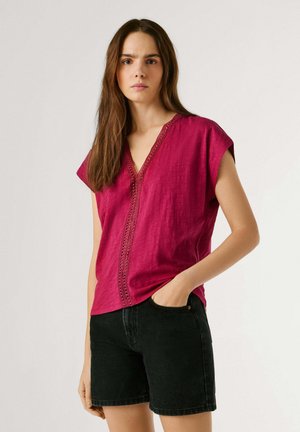 Pikkade pruunide juustega naine kannab sprossid kaunistatud magenta V-kaelusega pluusi ja musti teksapükse, seisab ühe käega taskus.