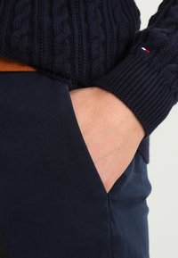 Donkerblauwe gebreide trui met kabelpatroon op de mouwen, gecombineerd met donkerblauwe broek met zijzak en een bruine leren riem.