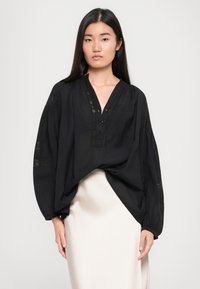 Sort oversized bluse med indsnørede manchetter, knapdetaljer og teksturerede accenter. V-udskæring og letvægtsstof, der hænger over en cremefarvet nederdel.