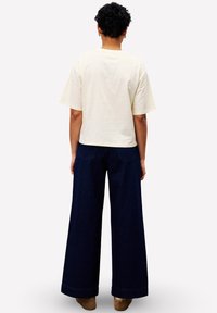 Persoon met kort, krullend haar die een losse crèmekleurige kortekorte mouw shirt, donkere, wijde broek en bruine schoenen draagt, staande met de rug naar ons gekeerd.