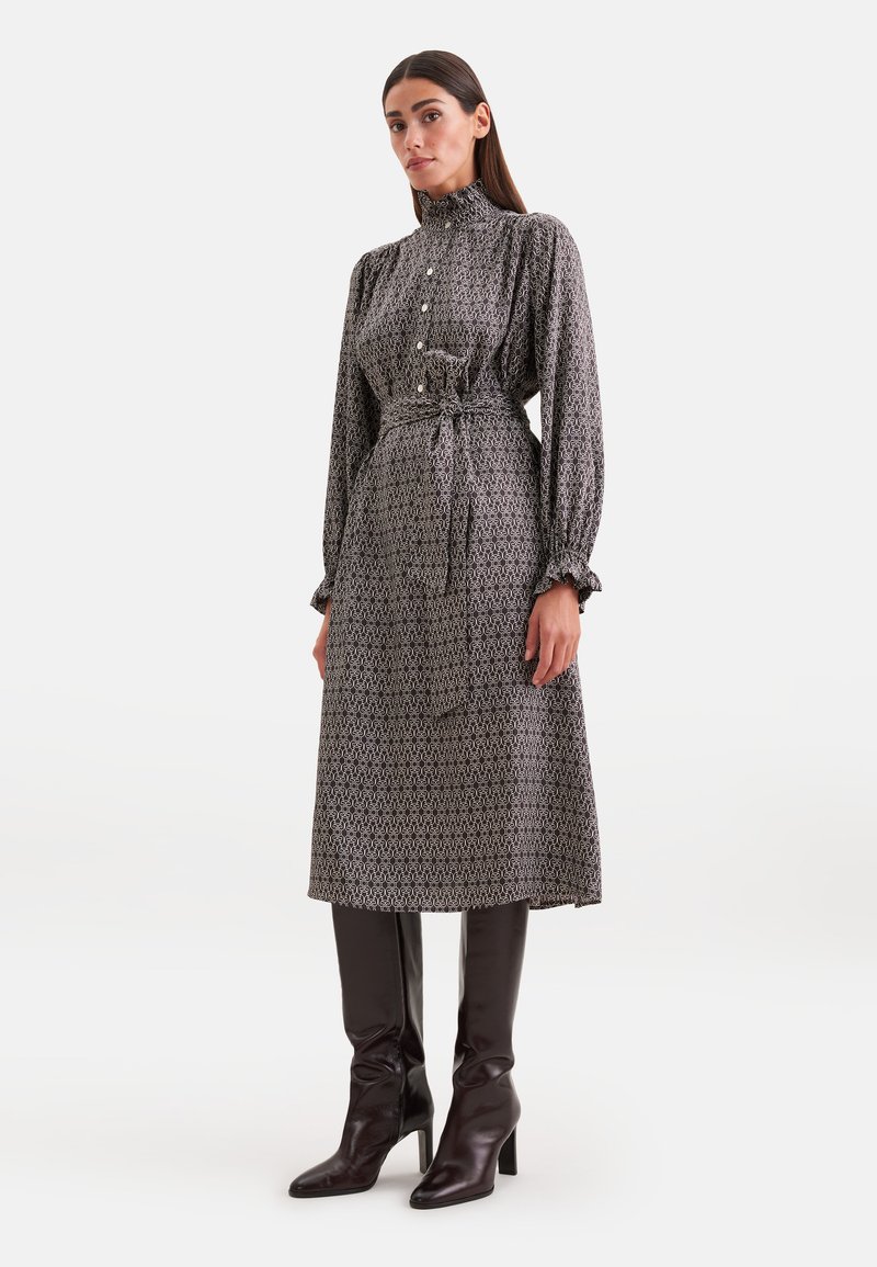 Femme debout portant une robe midi à manches longues à motifs, avec un col haut, une ceinture à la taille et des bottes noires à talons montant jusqu'au genou.
