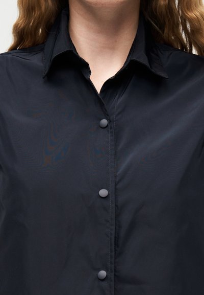 Femme aux longs cheveux ondulés portant une chemise noire à col boutonnée avec des boutons ronds noirs, visible du cou au milieu du torse.