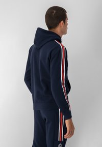 Sudadera con capucha de color azul marino en tejido suave, con detalles de franjas rojas y blancas a lo largo de las mangas, puños acanalados y un diseño ajustado.