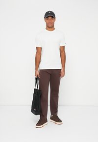 Camiseta blanca, pantalones marrones, zapatillas marrones con suelas blancas, bolsa tote negra. El modelo lleva una gorra oscura con un logo en la parte frontal.