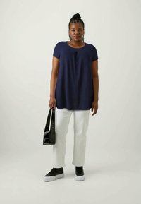 Námornícky modré voľné tričko s krátkymi rukávmi, skombinované s bielymi nohavicami a čiernymi teniskami. Drží čiernu kabelku na ramene.