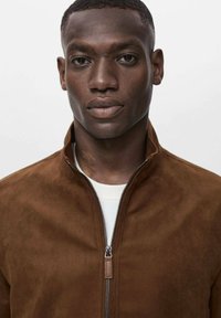 Chaqueta de ante marrón con collar alzado y cierre de cremallera plateada. Textura suave, diseño minimalista, combinada con una camiseta blanca por dentro.