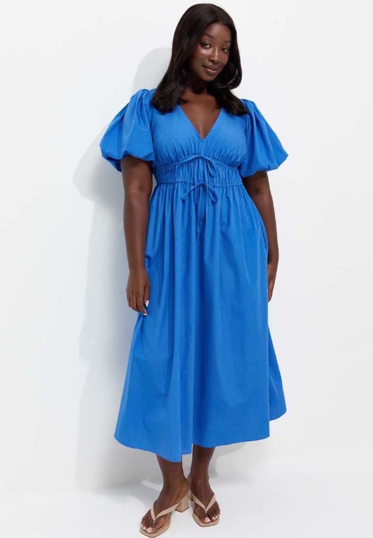 Woman with dark skin and hair wearing a bright blue midi dress with puff sleeves and beige heeled sandals standing against a white background.