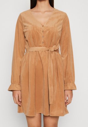 Kobieta ubrana w długą sukienkę z beżowego sztruksu z guzikami i przewiązanym paskiem w talii, stojąca na jednolitym tle.