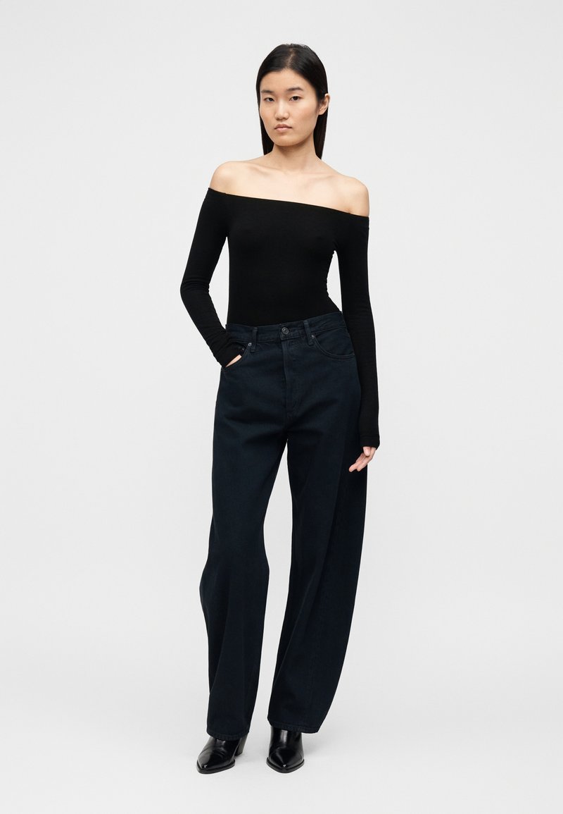 Haut noir à manches longues, décolleté épaules nues, associé à un jean en denim foncé taille haute et jambes larges, complété par des bottines noires à bouts pointus.