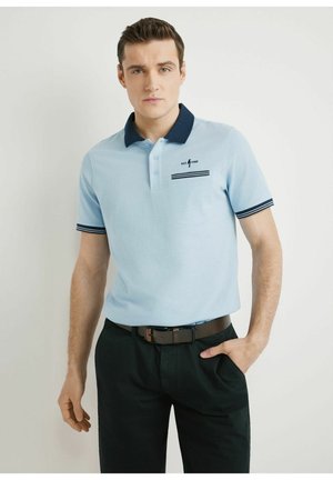 Mężczyzna ubrany w jasnoniebieską koszulkę polo z granatowym kołnierzem i paskami, czarne spodnie oraz czarny pasek, stojący na jednolitym tle.