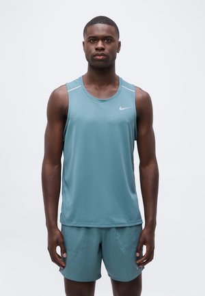 Hombre atlético de pie, quieto, vistiendo camiseta sin mangas Nike azul claro y pantalones cortos a juego, fondo blanco neutro.