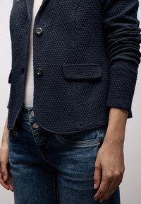 Gros plan sur une personne portant un blazer bleu marine texturé avec deux boutons noirs et un jean bleu, main détendue le long du corps.