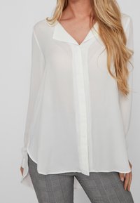 Blusa bianca a maniche lunghe realizzata in tessuto trasparente, con scollatura a V e chiusura con bottoni, abbinata a pantaloni grigi a scacchi.