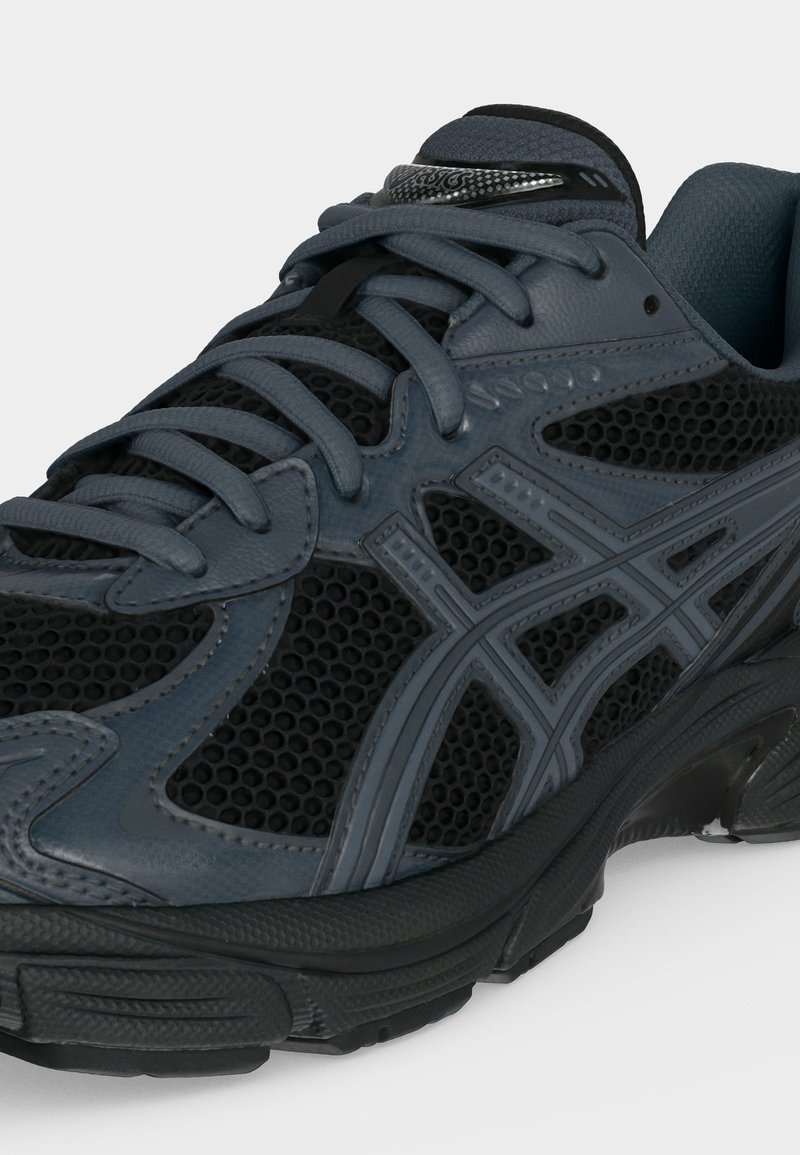 Zapatilla deportiva en gris oscuro y negro con tejido de malla, superposiciones sintéticas, cordones y suela de goma texturizada, mostrada desde un ángulo frontal lateral.