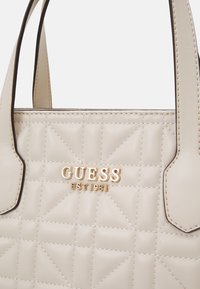 Beige gesteppte Handtasche mit goldenem "GUESS"-Logo und Nähdetails, ausgestattet mit zwei oberen Griffen und einem strukturierten Design.