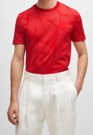 Mand iført en rød kortærmet mønstret t-shirt, der er stoppet ned i højtaljede hvide plisserede bukser, med hænderne afslappet langs siderne.