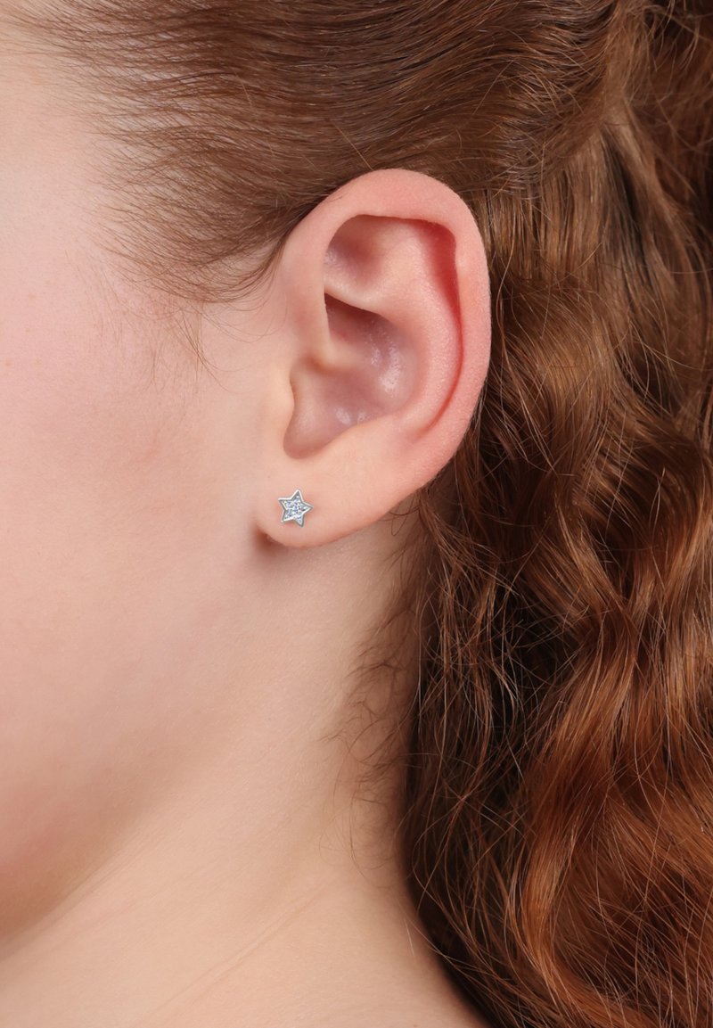 Pendiente de estrella plateada con un acento azul claro y un sutil brillo, llevado en el lóbulo de la oreja junto a un cabello rizado castaño.