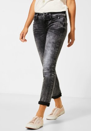 Persona con vaqueros negros descoloridos remangados y zapatillas rosas claras, caminando frente a un fondo claro uniforme.