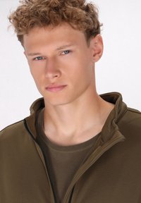 Olijfgroene zip-up jacket met een hoge kraag, gelaagd over een bruine t-shirt. Gladde textuur, aansluitend ontwerp en subtiele naaddetails.