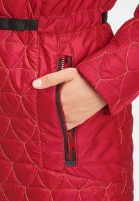 Rød quiltet jakke med guldstikninger, der har en lynlåslomme og et sort bælte. Glat tekstur med et geometrisk mønster på stoffet.