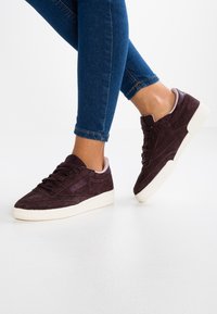 Mörklila mockaskor med vita gummisålar, med snörningsdesign och en mjuk rosa accent vid skaftet.