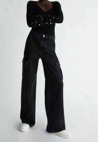 Pantalon cargo noir à jambes larges avec poches latérales, en denim, coutures détaillées et taille haute, associé à des baskets blanches.