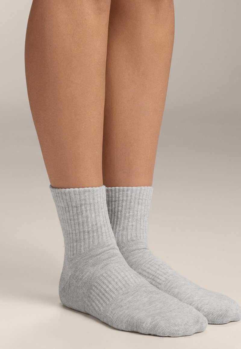 Beine, die schlichte hellgraue gerippte Bündchensocken tragen, stehen auf einer neutralen Fläche vor einem weichen beigen Hintergrund.