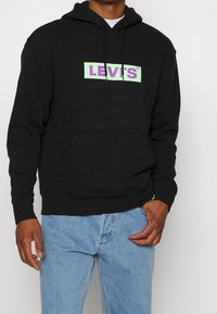 Sweat-shirt à capuche noir en coton, arborant un graphisme du logo Levi's en vert pastel et violet, avec une poche kangourou et des cordons de serrage.
