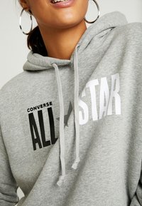 Felpa grigia realizzata in tessuto morbido, con la scritta "CONVERSE ALL STAR" in grassetto bianco e nero. Include dettagli con lacci.