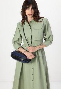 Rochie cu mânecă lungă, cu nasturi, dintr-un material verde deschis, având un buzunar pe piept; asortată cu o geantă din piele în două nuanțe, negru și bleumarin.