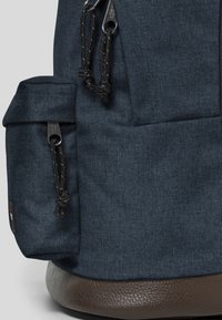 Mochila de tela azul oscuro con un acabado texturizado, que cuenta con un pequeño bolsillo lateral, cierre de cuerda y detalles en piel marrón en la base.