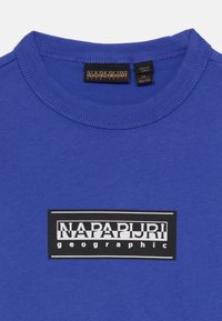 Zils kokvilnas T-krekliņš ar ribotu apaļu kaklu, ar melnu taisnstūrveida logotipu, kurā balto tekstu lasām "NAPAPIJRI geographic".
