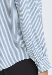 Blue and white vertically striped long-sleeve shirt cuff with white button, worn over black pants, showing side and lower sleeve details.