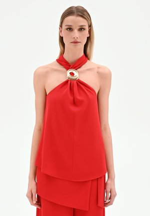 Femme aux cheveux blonds raides porte un top rouge dos nu avec un grand anneau doré au niveau de l'encolure, sur fond blanc.