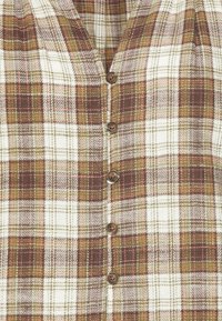Rutiga skjortan i kräm, brunt och grönt; har en V-ringning, vertikal knappknäppning och textil med en kontrasterande rutmönster.