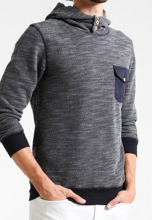 Mężczyzna w szarej dzianinowej bluzie z kapturem, ozdobionej trzema drewnianymi guzikami i granatową kieszenią, z rękami w kieszeniach białych spodni, na tle jednolitego białego tła.