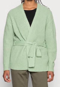 Jasnozielony sweter z dzianiny z paskiem w talii, z teksturowanym wzorem i otwartym przodem. Długie rękawy i ściągacze na mankietach.
